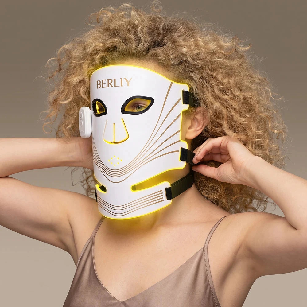 Woman adjusting the straps of a white BERLIY face and neck mask with yellow light visible at the edges.